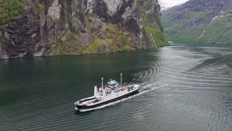 Car-and-tourist-ferry-full-of-people-cruising-in-spectacular-Geiranger-Fjord-Norway---Aerial-rotating-around-ship-showing-all-surrounding-landscape-while-keeping-ship-in-middle-of-frame