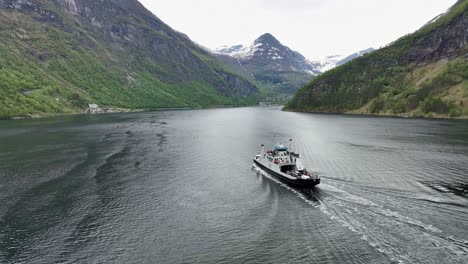 Following-tourist-ferry-route-from-Hellesylt-to-Geiranger-on-Geirangerfjord-with-Geiranger-village-in-background---Aerial-during-may-month-with-green-forest-and-snow-capped-mountains