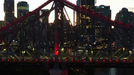 Story-bridge-adventure-climb,-tiny-people-climbing-on-iconic-cantilever-bridge-with-busy-traffic-crossing-the-river-at-dusk-and-illuminated-downtown-cityscape-of-Brisbane-city,-cinematic-aerial-shot