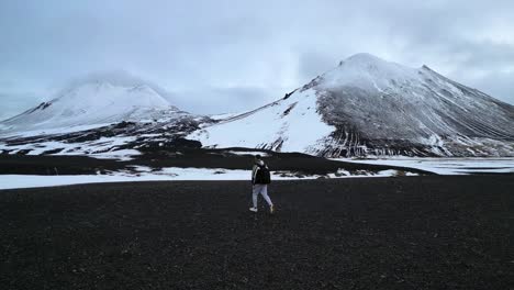 Drone-View-Following-Man-Walking-on-Black-Lava-Volcanic-Landscape-in-Iceland-on-a-Cloudy-Day-in-Wintertime
