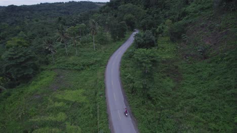 Drone-flies-over-a-small-road-with-motorbike-driving-at-Sumba-island,-aerial