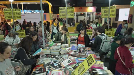 Tiro-Ascendente-Por-Encima-De-La-Gente-Mirando-Libros-Apilados-Desordenadamente-En-La-Mesa-De-Ofertas-Especiales