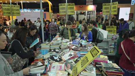 Tiro-Descendente-Sobre-Una-Mesa-De-Libros-Desordenada-Y-Ocupada-En-Una-Feria-Con-Multitudes-De-Personas-Caminando