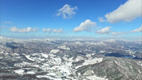 Eine-Drohnen-Luftaufnahme-Einer-Gefrorenen-Bergkette-Mit-Schnee-Und-Wolken-An-Einem-Strahlend-Blauen-Himmelstag