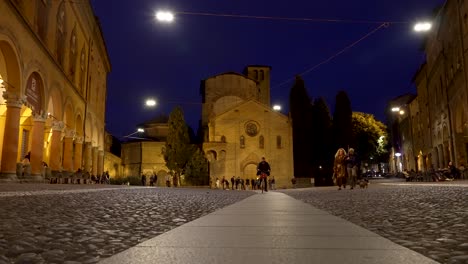 Eine-Aufnahme-Der-Von-Lichtern-Beleuchteten-Basilika-Von-Santo-Stefano-An-Einem-Schönen-Abend-Aus-Der-Tiefwinkelansicht,-An-Der-Ein-Paar-Vorbeigeht-Und-Einen-Entspannten-Spaziergang-Durch-Die-Nächtliche-Stadt-In-Bologna,-Italien,-Genießt