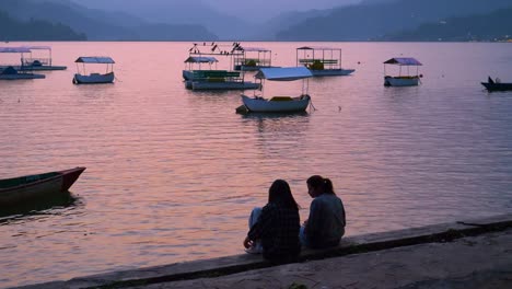 Two-women-sit-at-dock-of-pokhara-lake-talking-enjoying-beautiful-sunset