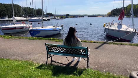 A-young-pregnant-woman-with-sunglasses-is-sitting-on-the-bench-in-a-harbor-with-a-view-of-small-yachts-on-a-sunny-calm-summer-day,-parallax-shot