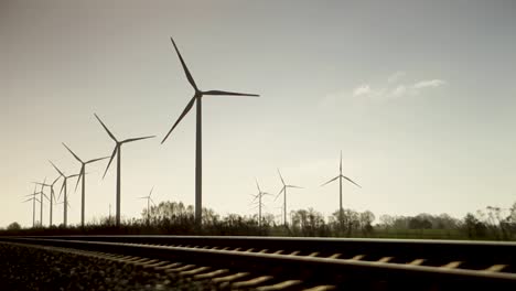 Panorama-view-spinning-wind-turbines-next-to-train-track,-sunset,-blue-sky