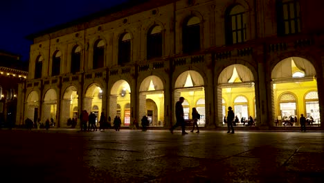 Lights-illuminating-the-arches-of-the-Palazzo-Re-Enzo-at-night,-as-people-walk-through-the-Piazza-Maggiore-on-a-beautiful-evening,-Bologna,-Italy
