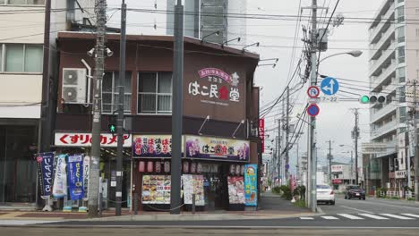 Street-Corner-In-Downtown-Hakodate-With-Businesses-With-Colourful-Shop-Front-Cars-Driving-Past