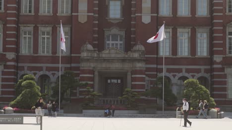 Vorderansicht-Des-Klassischen-Tokioter-Bahnhofsgebäudes-Mit-An-Den-Seiten-Wehenden-Japanischen-Flaggen-An-Einem-Sonnigen-Tag-Und-Herumlaufenden-Menschen