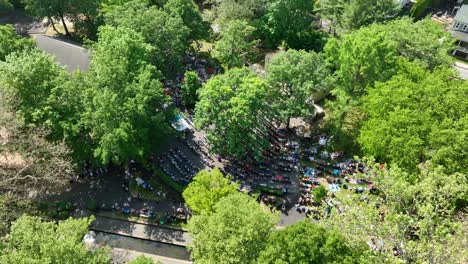 Panorama-Drohnenansicht-Eines-Großen-Parks-Mit-Mehreren-Gemeindemitgliedern,-Die-An-Einem-Klaren-Sommertag-Eine-Aufführung-Im-Amphitheater-Genießen