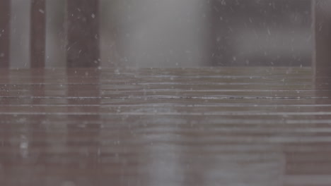 Close-up-shot-of-rain-drops-falling-on-a-wooden-table-making-splashes-with-chairs-in-the-background-in-slowmotion-LOG