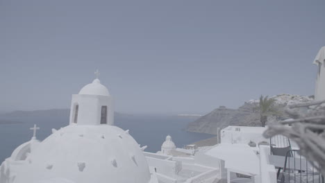 Überblick-über-Santorini,-Griechenland-An-Einem-Sonnigen-Tag,-Gesehen-Von-Einer-Terrasse-In-Zeitlupe-Und-Dem-Meer-Im-Hintergrund,-Das-Die-Sonne-In-Der-Nähe-Einer-Kirche-Oder-Eines-Gebäudes-Mit-Kreuzen-Auf-Dem-Dachbalken-Reflektiert