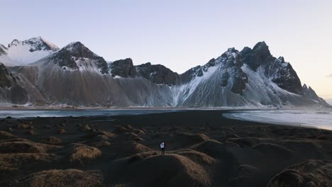 Vista-Aérea-De-Un-Hombre-Con-Una-Bandera-En-La-Playa-De-Stokksnes,-Frente-A-Las-Montañas,-Puesta-De-Sol-En-Islandia