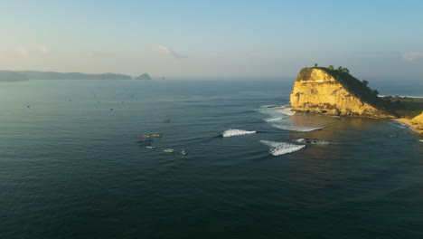 Panoramic-aerial-view-of-Gerupuk-surf-spot-with-surfers-riding-the-wave,-cliffs-in-the-middle-of-the-Bali-sea