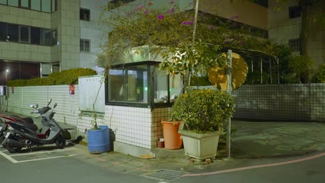Security-guard-shack-covered-in-plants-next-to-parked-mopeds-at-night-in-taiwan