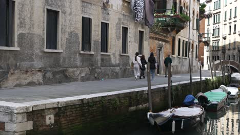 Touristen,-Die-Am-Kanal-Von-Venedig-Entlang-Spazieren,-Mit-Einer-Reihe-Kleiner-Leerer-Boote-Vor-Anker