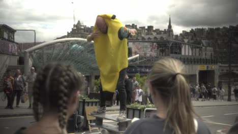 Witness-an-extraordinary-man-performing-a-captivating-balancing-act-by-jumping-onto-wooden-plank-in-the-vibrant-city-of-Edinburgh,-captivating-a-crowd-as-two-girls-watch-in-the-foreground