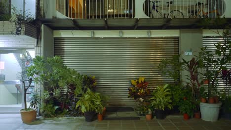 Metal-grated-front-door-garage-entrance-with-potted-tropical-plants-at-night-in-asia