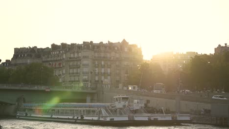 Tour-boat-barges-on-the-Seine-river-as-the-sun-sets-behind-buildings,-Pan-right-shot