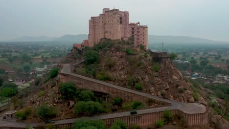 aerial-drone-camera-moving-forward-showing-Leela-Bisangarh-Jaipur-fort-with-only-one-way-to-go-up-or-down-with-vehicles-passing-by-and-core-residential-houses-surrounding-the-fort