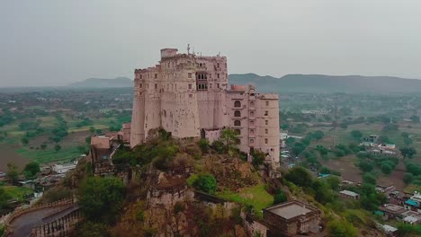 aerial-drone-camera-zoom-into-the-showing-Leela-Bisangarh-Jaipur-fort-with-only-one-way-to-go-up-or-down-with-vehicles-passing-by-and-core-residential-houses-surrounding-the-fort