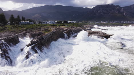 Angry-ocean-wave-crash-into-rocky-shoreline-creating-big-splash,-Hermanus-aerial