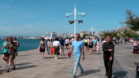 Slow-motion-shot-of-people-walking-on-promenade-in-Santander-City-against-blue-sky-in-summer---In-background-luxury-cruiser-ship-at-port