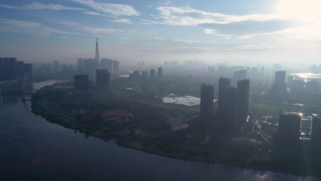 Fly-in-drone-shot-over-Saigon-River-towards-new-residential-developments-in-Thu-Thiem-on-misty,-sunny-morning