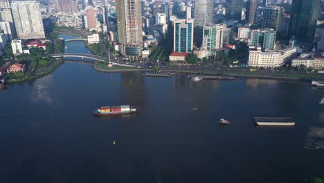 Drone-shot-over-Saigon-River-with-working-container-and-freight-boats-and-Ho-Chi-Minh-City-skyline-on-a-sunny,-clear-morning