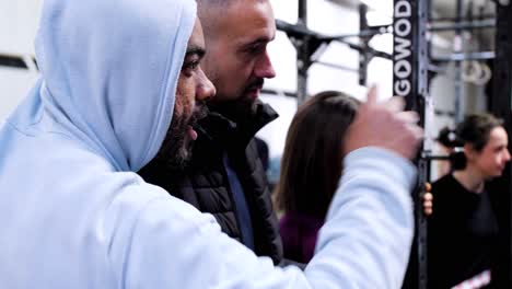 Amigos-Observando-Una-Competencia-De-Crossfit-Debajo-De-Las-Plataformas:-Uno-Sostiene-Un-Café-Mientras-El-Otro-Explica-Un-Movimiento,-Mientras-La-Mujer-Morena-Escucha