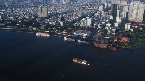 Fly-in-drone-shot-over-Saigon-River-waterfront-with-working-boats-on-the-river-and-tour-boats-at-the-wharf-on-a-sunny,-clear-morning