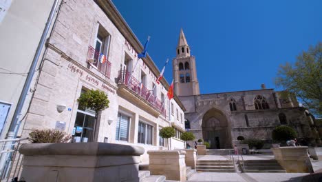Día-Soleado-En-La-Plaza-Del-Ayuntamiento-De-Montagnac:-Banderas-Francesas,-Europeas-Y-Occitanas-Ondeando,-Con-La-Iglesia-Del-Pueblo-Al-Fondo