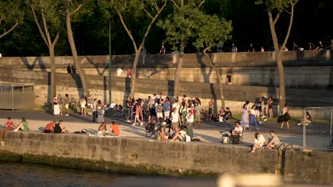 Gente-En-La-Orilla-Del-Río-Sena-Disfrutando-De-Una-Puesta-De-Sol-Vista-Desde-Un-Barco-Fluvial,-Tiro-Panorámico-A-La-Izquierda