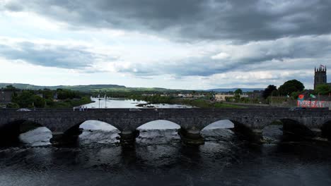 Drone-flying-towards-Thomond-Bridge-across-Shannon-River-on-overcast-day,-Limerick