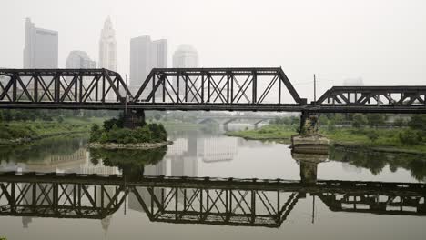 Brücke-Für-Züge-über-Einen-Fluss-Mit-Der-Skyline-Von-Columbus-Ohio-An-Einem-Nebligen,-Rauchigen-Tag