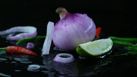 Slow-motion-green-chilli-and-chopped-rings-falling-onto-lemon-lime-and-onion-with-rippling-water-surface-against-black-background