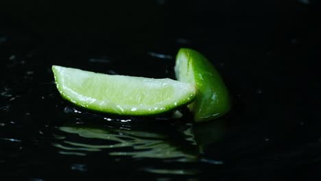 Slow-motion-fresh-segment-of-green-lime-falling-onto-rippling-water-surface-isolated-on-black,-Cooking-concept