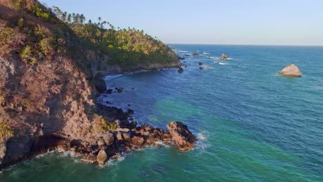 Aerial-establishing-drone-flight-along-coastline-with-growing-palm-trees-and-waves-of-Caribbean-sea-at-sunset