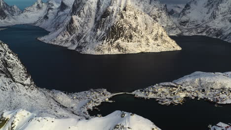 Aerial-view-tilting-over-the-snowy-Reinebringen-peak,-revealing-the-sunlit-Olstinden-mountain,-in-Reine,-Lofoten,-Norway