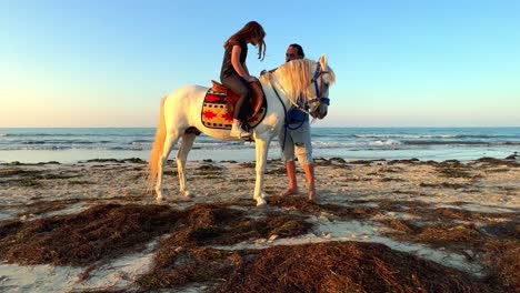 Bearded-man-instruct-little-girl-with-red-hair-on-white-horse-before-riding-on-beach-along-sea-shore-at-sunset