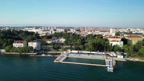 Panorama-Costero-Desde-Arriba-Con-Piscina-Marina-Y-Plataforma-De-Buceo,-Nadadores-Y-Ciudad-Zadar,-Croacia
