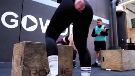 In-CrossFit,-a-girl-is-jumping-onto-a-box-while-her-trainer-and-others-watch-from-the-side