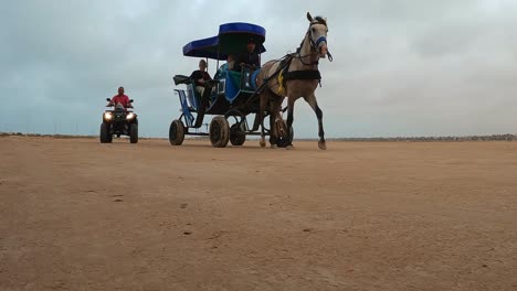 Vorgespannte-Pferdekutsche-Mit-Touristen-Entlang-Der-Trockenen-Wüste-Tunesiens,-Hinter-Dem-Ein-Quad-Fährt