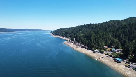 Aerial-drone-flight-over-Twanoh-State-Park-Lake,-Washington,-showcasing-the-tranquil-landscape,-clear-blue-skies,-and-green-pine-trees-along-the-coastline