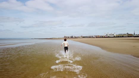 A-man-with-no-shirt-running-fast-through-sea-water-on-a-beach,-aerial
