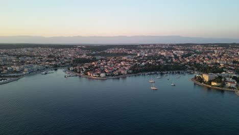 An-aerial-panorama-following-coastline-in-Zadar-city-in-Croatia,with-blue-Adriatic-Sea,-sailboats,-marina,-houses-and-buildings-and-distant-mountain-Velebit-at-sunset