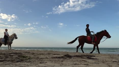 Low-angle-of-people-riding-horses-at-sunset-on-seashore-along-sandy-beach