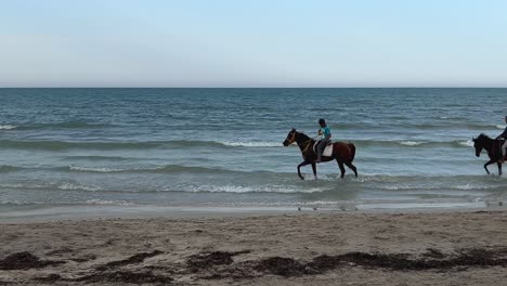 Menschen-Reiten-Auf-Zwei-Pferden-Entlang-Der-Küste-Im-Meerwasser